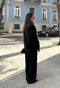 Frau im schwarzen Anzug und Sonnenbrille steht auf dem Bürgersteig nahe einem Zebrastreifen, im Hintergrund ein blaues Gebäude und ein schwarzes Auto.