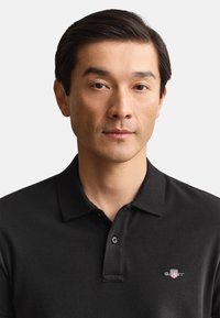 Man with short dark hair wearing a black polo shirt with a small logo on the chest, neutral expression, white background.