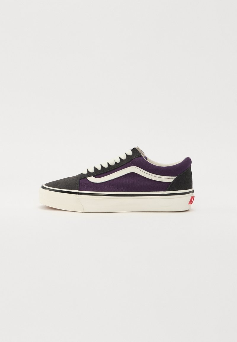 Zapatilla de cordones con un upper de lona negro y morado, suela de goma blanca y una franja de logo en el lateral. Presenta una puntera y un talón en negro contrastante.