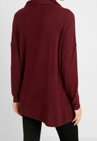 Pullover bordeaux a maniche lunghe con collo a scialle, texture morbida, vestibilità ampia e lunghezza fino a metà fianchi, abbinato a leggings neri.