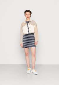 Jaqueta de ganga bege e creme, cortada, sobre um vestido listrado em azul e branco. A modelo usa ténis brancos com atacadores laranja; pose minimalista.