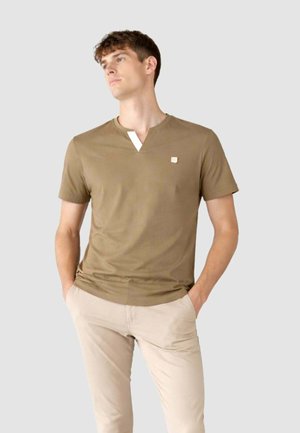 Giovane uomo che indossa una maglietta marrone a maniche corte con scollo a V e pantaloni beige chiaro, in piedi con le mani nelle tasche, che guarda leggermente di lato.