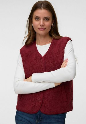 Jeune femme aux longs cheveux bruns porte un gilet polaire bordeaux confortable sur une chemise blanche à manches longues, debout les bras croisés.