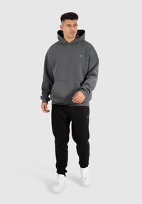 Grauer Kapuzenpullover aus weichem Material mit einer Fronttasche und einem Kordelzug an der Kapuze, kombiniert mit schwarzen Jogginghosen und weißen Sneakers.
