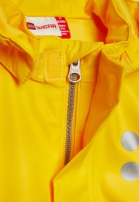 Yellow waterproof jacket with a front zipper, featuring a YKK fastener and silver dot accents. Interior tag includes size and care information.