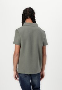 Kurzarm-Poloshirt aus strukturiertem olivgrünem Stoff, mit spitzem Kragen und geradem Saum, getragen mit blauen Jeans.