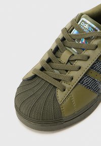 Zielony sneaker z teksturowanym gumowym noskiem, siateczkową boczną panelem i oliwkowymi skórzanymi wierzchami. Posiada czarne sznurowadła i markową holograficzną metkę.