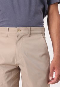Pantalons beiges avec fermeture éclair et bouton, dotés de poches avant et d'une texture lisse. Portés avec un T-shirt gris.