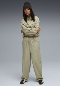 Veste de sport vert olive clair avec des poignets élastiques et un pantalon ample assorti. Logo Puma noir sur la blouse et le pantalon. Bonnet en tricot marron.