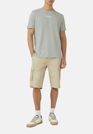 Mann trägt hellgraues T-Shirt mit kurzen Ärmeln, beige Cargoshorts, weiße Socken und beige Turnschuhe, steht mit den Händen in den Taschen.