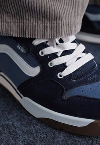 Mornarsko plavi Vans tenisice s bijelim vezicama, od suđene i mrežaste tkanine, s bijelim bočnim prugom. Imaju bijelu srednju podlogu i smeđu vanjsku podlogu.