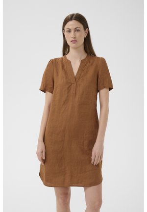 Kobieta stojąca, ubrana w brązową, z rudawym odcieniem lnianą sukienkę do kolan z krótkim rękawem, rozciętym dekoltem i kolczykami kołami na jednolitym tle.