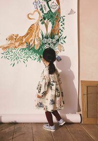 Kind in einem blumengemusterten cremefarbenen Kleid mit Puffärmeln, dunkelroten Strumpfhosen und hellblauen Schuhen, bewundert ein lebhaftes Wandgemälde, das einen Tiger zeigt.