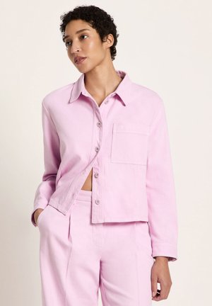 Woman with short curly hair wearing a light pink button-up jacket and matching high-waisted pants, standing with one hand in pocket.