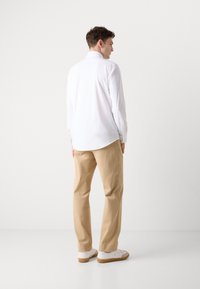 Hvit langermet skjorte med en svakt buet hem, kombinert med lyse beige bukser og hvite joggesko, vist på en modell sett bakfra.