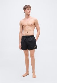 Μαύρο μαγιό σορτς με κορδόνι στη μέση, πλαϊνές τσέπες και χρυσή διακριτική λεπτομέρεια με λογότυπο. Υλικό από πολυεστέρα, σχέδιο μέχρι το γόνατο.