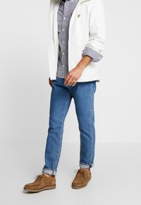 Giacca bianca con cappuccio, camicia a quadri, jeans di denim blu e scarpe in suede marroni. Caratteristiche notevoli includono il motivo a quadri e i polsini arrotolati.