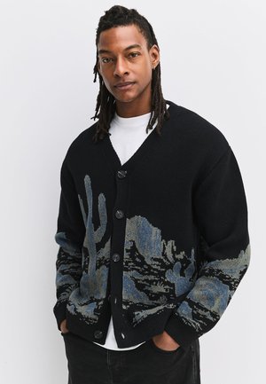 Man with dreadlocks wearing black cardigan with blue-gray cactus and desert landscape pattern, hands in pockets, standing against white background.