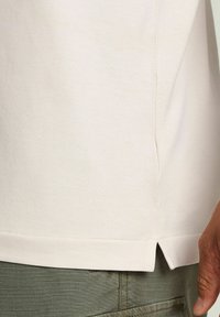 Polo shirt beige chiaro in tessuto testurizzato. Presenta un orlo spaccato e maniche strette, abbinato a pantaloni verde oliva.