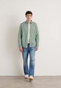 Lys grønn skjorte med knapper over en hvit t-skjorte, kombinert med blå jeans og hvite joggesko. Glatt stoff, avslappet passform, uten mønstre.