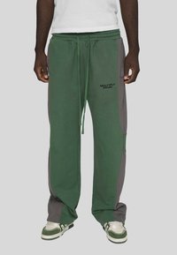 Bărbat îmbrăcat în pantaloni de trening largi verzi și gri cu logo-ul "MULTIPLY APPAREL" și adidași alb-verzi pe un fundal gri.