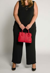 Kobieta ubrana w czarny bezrękawnik i spodnie, trzymająca czerwoną torebkę obiema rękami na wysokości brzucha, ubrana w czarne buty i biżuterię.