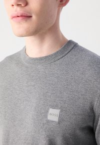 Nahaufnahme einer Person, die einen grauen Pullover trägt, auf dessen Brust ein kleines quadratisches Logo-Patch mit der Aufschrift "BOSS" zu sehen ist.