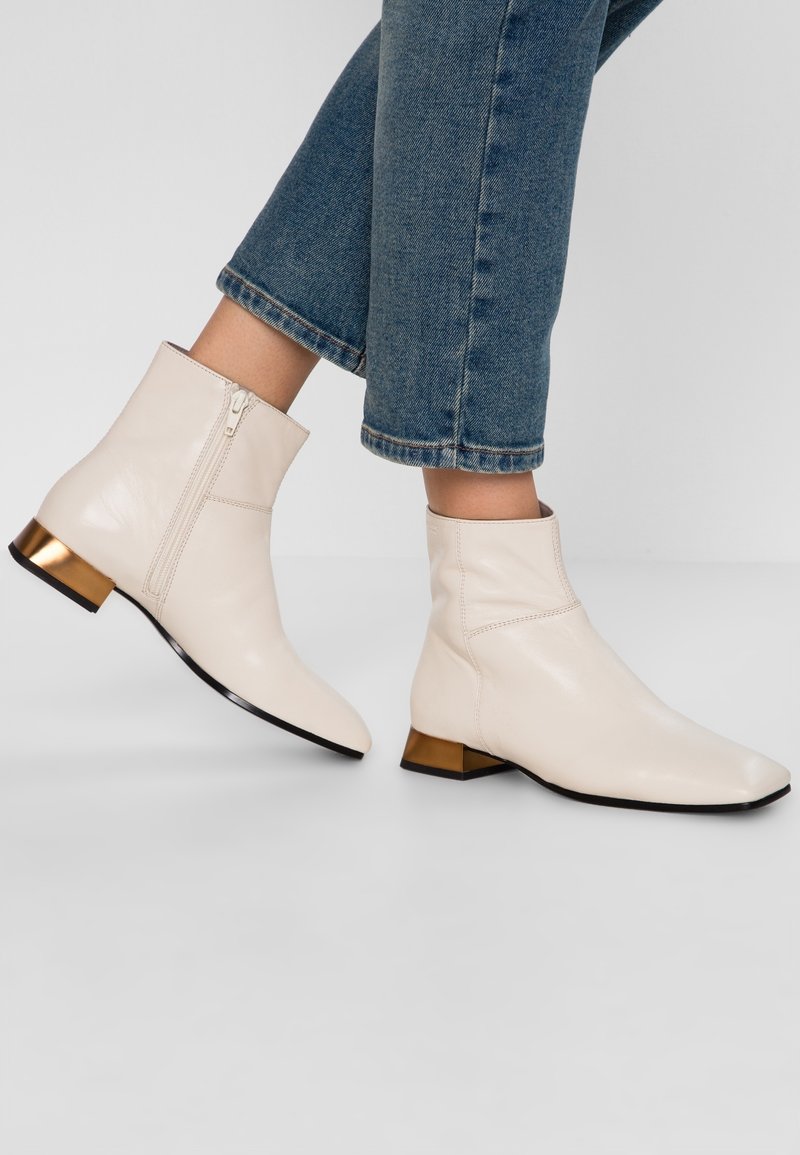 Bottes en cuir crème montant à la cheville avec fermetures éclair latérales, talons carrés métalliques dorés bas, portées avec un jean bleu délavé raccourci sur un fond clair.