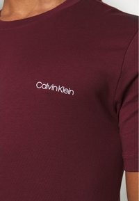 T-shirt bordeaux a manica corta con scollo rotondo, realizzato in cotone elasticizzato, che presenta il logo bianco "Calvin Klein" sul lato sinistro del petto.