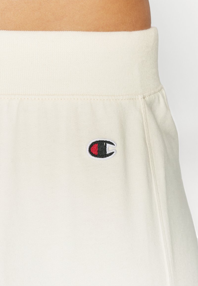 Pantalones de chándal color crema con una cintura elástica y un pequeño logo bordado en una combinación de colores negro, rojo y blanco.