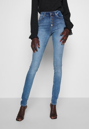 Modelka nosi jasnoniebieskie obcisłe jeansy z wysokim stanem i zamknięciem na pięć guzików, zestawione z czarnym topem z długim rękawem oraz czarnymi sandałami na obcasie z odkrytymi palcami.