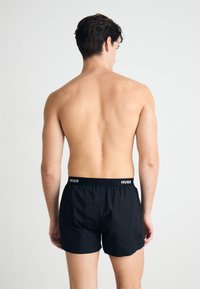 Boxeri din bumbac negri, cu talie elastică, având un logo alb "HUGO". Modelul este prezentat din spate, fără tricou.