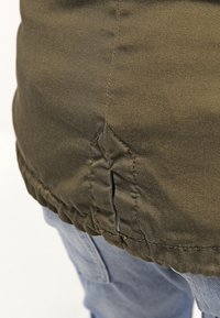 Gros plan sur l'ourlet d'une veste vert olive avec une fente latérale sur un jean en denim bleu clair.