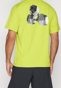 Hombre con camiseta verde lima con un gráfico en blanco y negro de tres pesas en la parte superior de la espalda, combinada con pantalones cortos negros.