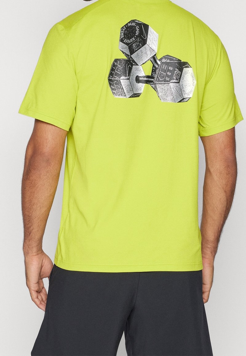 Hombre con camiseta verde lima con un gráfico en blanco y negro de tres pesas en la parte superior de la espalda, combinada con pantalones cortos negros.