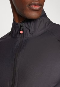 Schwarze Sportjacke mit hohem Kragen, strukturiertem Stoff, vorderem Reißverschluss mit rotem Zugband und tonalem Nähen für Details.