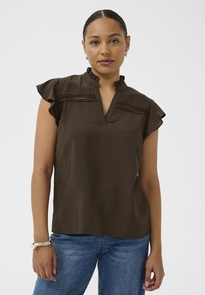 Femme aux cheveux tirés en arrière, portant un chemisier sans manches brun foncé avec des détails en dentelle et un jean bleu, debout devant un fond uni.