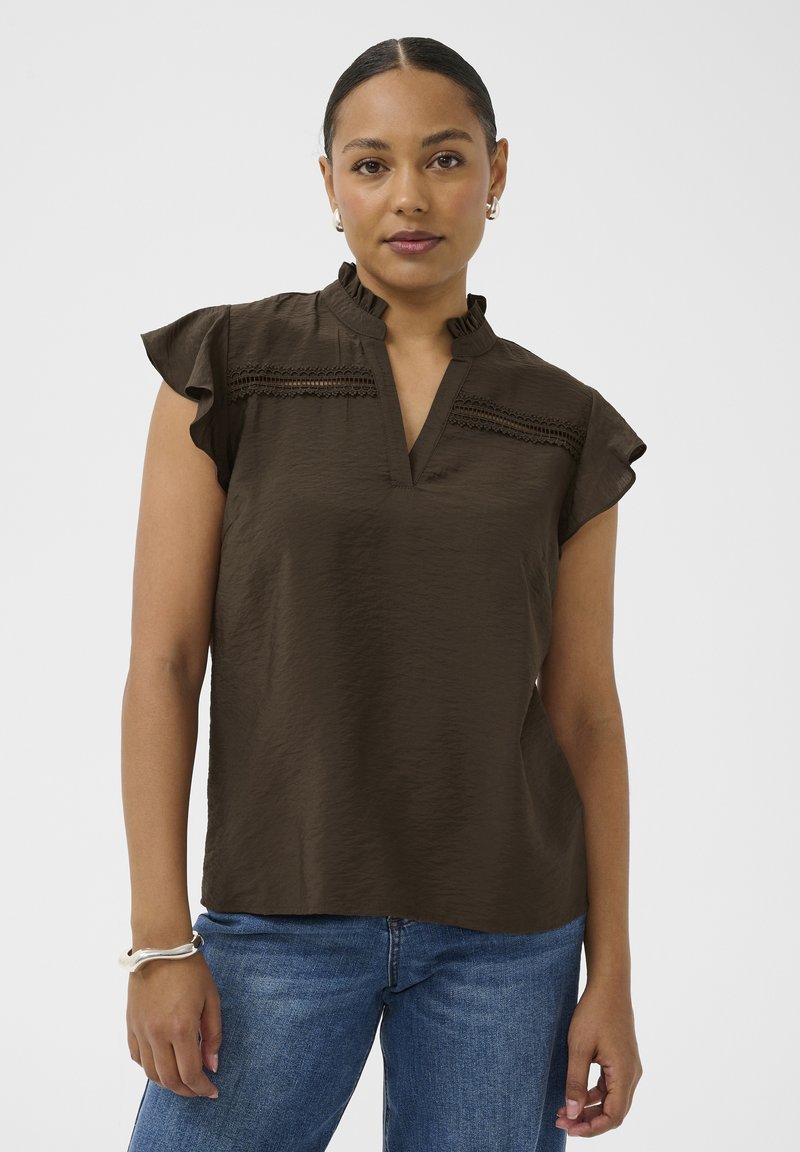 Femme aux cheveux tirés en arrière, portant un chemisier sans manches brun foncé avec des détails en dentelle et un jean bleu, debout devant un fond uni.