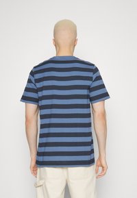 Person med kort blont hår iförd en blå och svart horisontellt randig t-shirt och off-white byxor, sedd bakifrån.