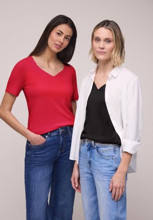 Dwie kobiety stoją obok siebie; jedna ma na sobie czerwony top z dekoltem w serek i dżinsy, druga czarny top, białą koszulę i jasnoniebieskie dżinsy.