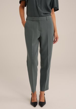Femme portant un pantalon gris ajusté et des talons noirs pointus, debout devant un fond uni.
