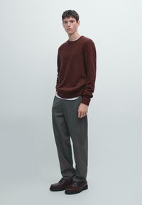 Maglione a maglia color mogano, pantaloni grigi, scarpe stringate marroni. Il maglione presenta un collo rotondo e un orlo a coste. I pantaloni sono sartoriali e a gamba dritta.