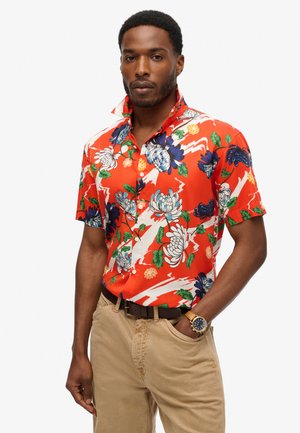 Camisa corta de manga roja con estampado floral que presenta grandes flores azules y blancas, hojas verdes y un cuello abotonado. Combinada con pantalones beiges.