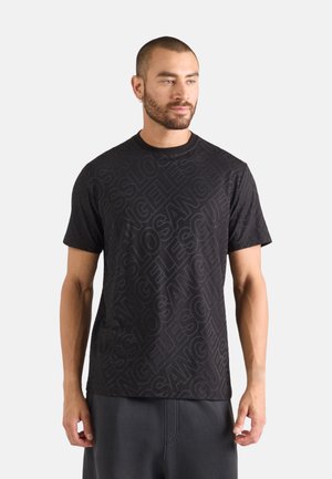 Homme aux cheveux courts et à la barbe, portant un t-shirt noir avec le texte répété « Los Angeles Guess » disposé en motif diagonal.