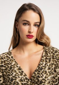 Šaty s leopardím vzorom v odtieňoch zlatej a hnedej, s hlbokým výstrihom do V a prúdiacou tkaninou. Dlhé zlaté náušnice dotvárajú vzhľad.