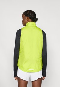 Ανοιχτό πράσινο ελαφρύ γιλέκο με ψηλό γιακά, κατασκευασμένο από λείο, λαμπερό ύφασμα, συνδυασμένο με μαύρο μακρυμάνικο πάνω μέρος και λευκά πλισέ σορτς.