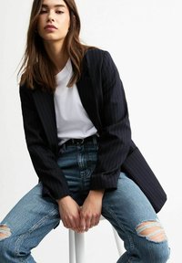 Zwarte pinstripe blazer over een witte t-shirt, gecombineerd met verouderde lichtblauwe jeans. De jeans hebben grote scheuren en een comfortabele pasvorm.