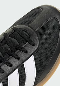Black athletic shoe featuring a textured leather upper with mesh sections, black laces, and three white stripes along the side. Brown rubber sole.
