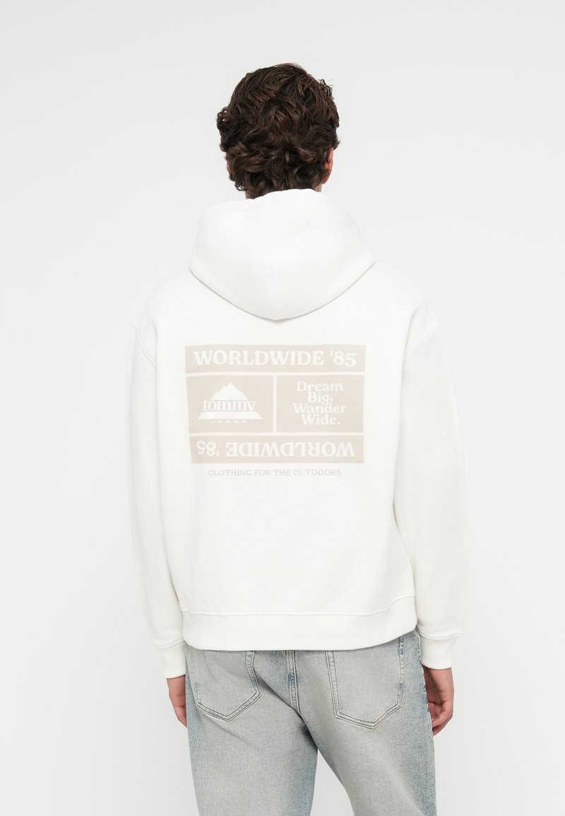 Λευκή hoodie με μια μεγάλη μπεζ γραφιστική στην πλάτη, που εμφανίζει το κείμενο "WORLDWIDE '85" και "Dream Big. Wander Wide." Διαθέτει αξιοσημείωτους ριγωτούς μανσέτες.