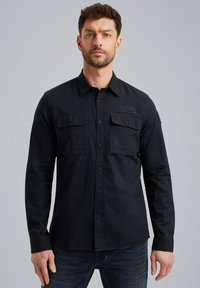 Camisa negra de manga larga con cierre de botones en el frente, dos bolsillos en el pecho y un parche texturizado en el hombro. Hecha de tejido ligero.